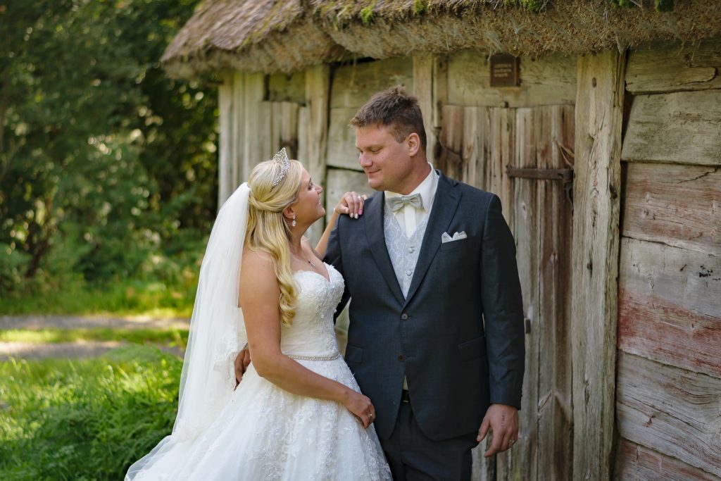 Brud håller hand på brudgummens axel i en glittrig brudklänning från Demetrios. Bruden har slöja i håret under bröllopsfotograferingen. De står vid ett ödehus i en hembydspark för unika bröllopsfoton.