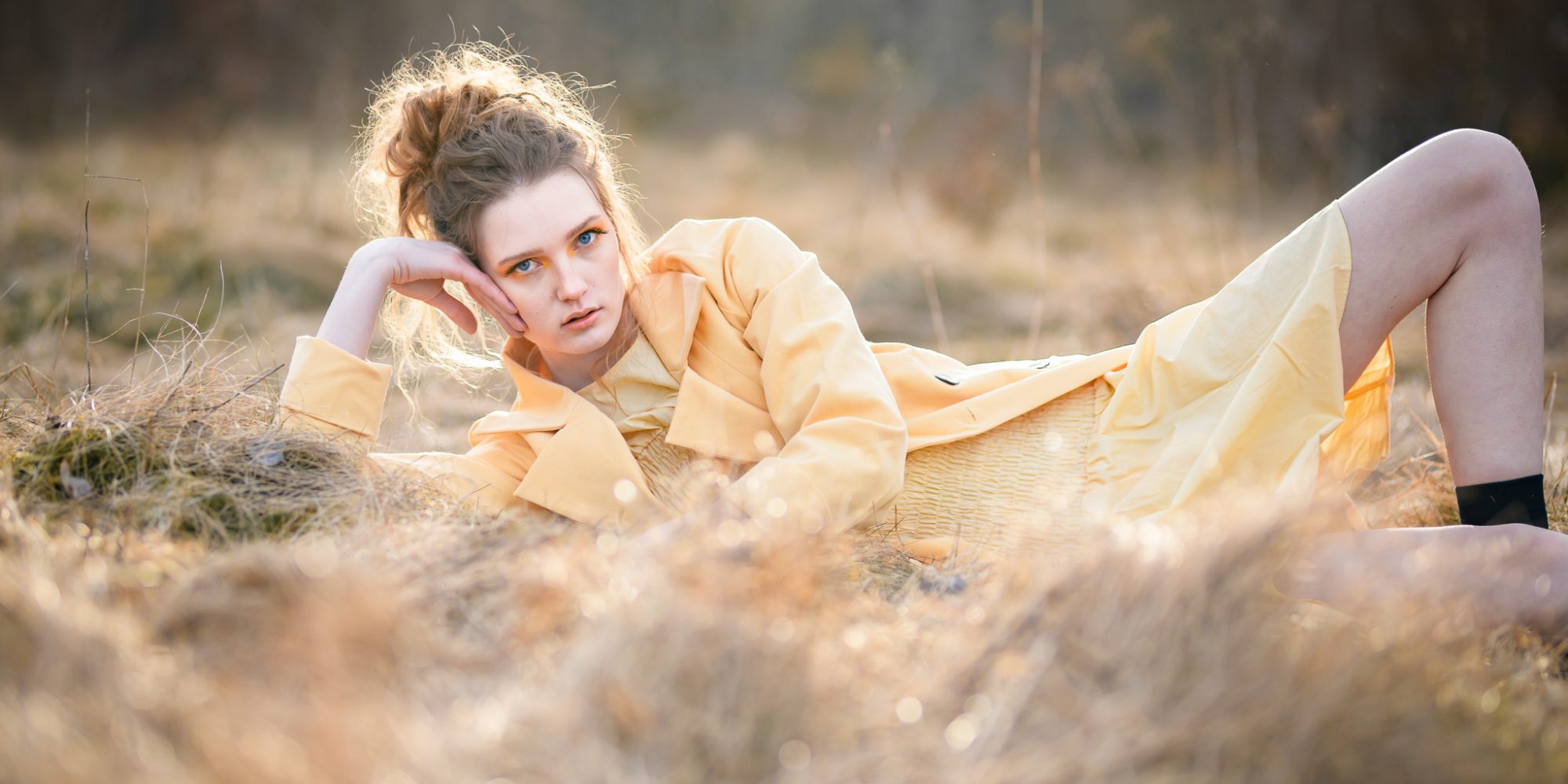Egoboost-porträtt, Modefotografering. Modell ligger i gul rock på en äng med dött gräs i en kaxig pose.